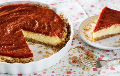 Fotografia em tons de vermelho em uma mesa com uma toalha branca com flores coloridas, um recipiente redondo raso com a cheesecake dentro dele. Ao lado, um prato raso branco com uma fatia da cheesecake.