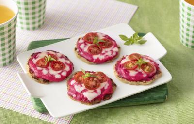 Foto da receita de mini pizza. Observa-se uma bandeja branca com 4 mini pizzas em cima, com recheio de creme rosa de beterraba, tomates cereja e queijo derretido, decoradas com folha de manjericão. O fundo é verde claro.