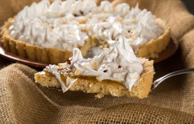 Fotografia de uma espátula segurando uma fatia de torta de cupuaçu e castanha, com merengue por cima. Ao fundo, a mesma torta, inteira sobre apoio redondo na cor marrom.