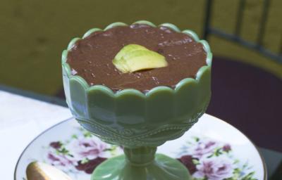 Fotografia em tons de verde em uma mesa de madeira com uma toalha branca de renda, um prato redondo branco com flores roxas e folhas verdes, uma taça verde com a mousse de abacate com chocolate dentro dela.