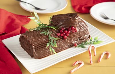 Fotografia em tons de vermelho em uma bancada de madeira amarela, um pano vermelho ao lado e ao centro, um recipiente retangular branco com o rocambole de chocolate em cima com decoração natalina.