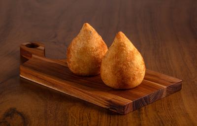 Fotografia de duas coxinhas de frango sobre uma tábua pequena de madeira. A tábua está sobre uma mesa de madeira.