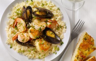 Fotografia em tons de branco e bege de uma bancada branca vista de cima. Contém um prato redondo branco com risoto com mariscos e ao lado um garfo para servir.