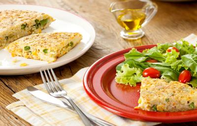 Fotografia em tons de vermelho e amarelo em uma bancada de madeira com um prato branco raso redondo ao fundo com a tortilla de arroz e ao lado, um prato vermelho com uma fatia da tortilla e salada de alface e tomate cereja.