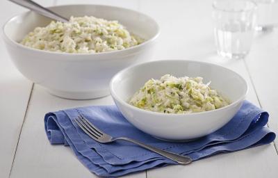 Fotografia em tons de branco e azul de uma bancada de madeira, sobre ela um paninho azul com um recipiente redondo com o arroz. Ao fundo um recipiente redondo com arroz, uma colher e um copo de vidro com água.