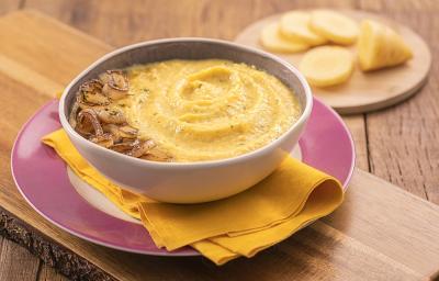 Foto em tons de amarelo da receita de creme de mandioquinha com cebola dourada servida em um bowl de porcelana branco sobre pano amarelo em cima de um prato com borda cor de rosa. Esses elementos sobre uma tábua de madeira e ao fundo mandioquinha cortada