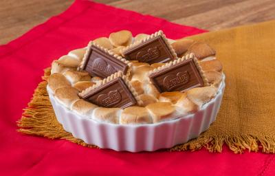 Fotografia de uma forma branca de vidro com s'mores que foi ao forno, e por cima, quatro biscoitos de chocobiscuit. A forma está apoiada sobre um paninho de tom marrom.