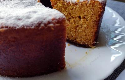 Fotografia em tons de bege em uma bancada com toalha cinza, um prato branco redondo raso, um bolo de milho cortado ao meio com açúcar polvilhado em cima.