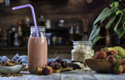 Fotografia de uma bebida densa de morango com leite MOÇA e neston que está em uma garrafa de vidro decorativa, com um canudo roxo e frutas no fundo.