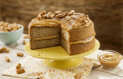 Fotografia em tons de amarelo em uma mesa de madeira com um pano bege com flores laranja e amarela, um suporte amarelo com o bolo de Moça Churros. Ao lado, um potinho com doce de leite. Ao fundo, um potinho azul com churros pequenos.