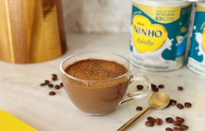 Fotografia em tons de amarelo e azul em uma bancada de mármore com uma xícara de vidro ao centro e o mocaccino dentro dela. Ao fundo, latas de Ninho Adulto e alguns grãos de café espalhados.