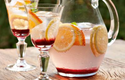 Fotografia em tons de rosa em uma bancada de madeira clara com uma jarra de vidro com o refresco de hibisco e frutas cítricas dentro, decorando com rodelas de laranja. Ao lado, duas taças de vidro com o refresco.