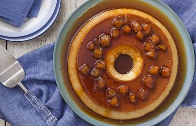 Fotografia em tons de caramelo e azul de uma mesa de madeira vista de cima, ao centro um recipiente redondo azul por cima de um tecido azul, dentro dele está o pudim com a calda de caramelo por cima e decorado com bananas carameladas.