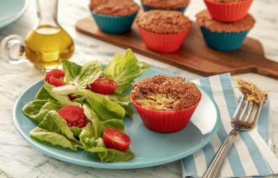 Fotografia em tons de verde em uma bancada de madeira de cor branca. Ao centro, um prato redondo azul contendo 1 muffin com um garfo ao lado. Ao fundo, uma tábua de madeira com mais muffins e um recipiente com azeite.