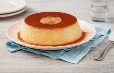 Fotografia em tons de caramelo, bege e azul claro. Ao centro até o pudim com calda de caramelo em cima de um prato branco e um guardanapo de tecido azul claro e uma espátula para servir. Fotografia em tons de caramelo, bege e azul claro. Ao centro até o pudim com calda de caramelo em cima de um prato branco e um guardanapo de tecido azul claro e uma espátula para servir.