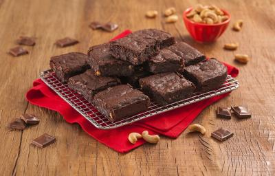 Fotografia em tons de marrom e vermelho de uma bancada de madeira com um paninho vermelho, sobre ele um prato retangular com pedaços de brownie. Ao fundo um recipiente redondo com castanha-de-caju.