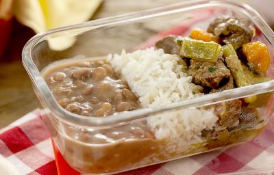 Fotografia em tons variados, com pote de vidro ao centro com feijão, arroz e picadinho de carne com legumes, sobre guardanapo quadriculado em branco e vermelho, ladeado por três panelas e uma ponta de pano amarelo ao fundo sobre bancada de madeira.