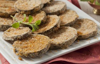 Fotografia em tons de vermelho em uma bancada de madeira com um pano vermelho, um recipiente branco retangular com as berinjelas à milanesa prontas.