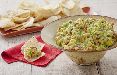 Fotografia em tons de verde e vermelho em uma bancada de madeira com um guardanapo de papel vermelho e sobre ele nachos e guacamole. Ao lado, um recipiente grande e redondo em formato de chapéu bege com o guacamole dentro dele.