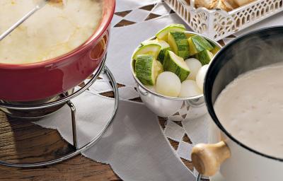 recipientes nas cores vermelho e branco ambos comporta o fondue de queijo, outros recipientes estão com os acompanhamentos para o fondue como pão, presunto entre outros.