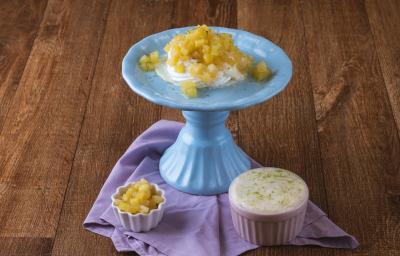 Foto da receita de pavlova de limão com abacaxi, servida em um prato azul, com creme ao lado em um pote rosa e outro pote branco com pedaços de abacaxi, tudo em uma bancada de madeira com um pano em tom violeta