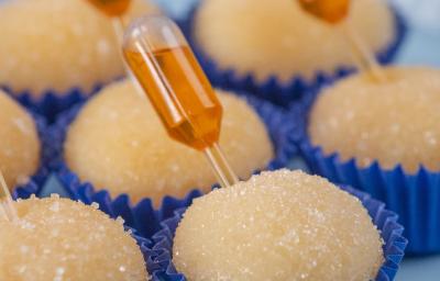 Fotografia de sete forminhas azuis juntas com brigadeiro de pudim, em cada unidade tem uma mini pipeta espetada com calda de caramelo. Os brigadeiros estão apoiados em um apoio decorativo azul claro.