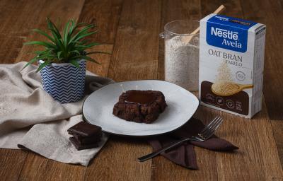 Fotografia de um pedaço de brownie com um quadrado de chocolate ao centro, sobre um prato branco com borda preta. O brownie está ao lado de uma embalagem de aveia farelo, sobre uma mesa de madeira.