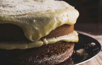 Fotografia de um bolo de abóbora com creme de coco por cima. O bolo está recheado com o mesmo creme, e está sobre um prato raso preto.