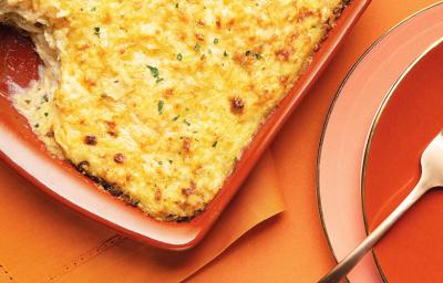 Fotografia em tons dourado e ferrugem, com refratário de cor ferrugem ao centro com escondidinho de bacalhau gratinado salpicado de cheiro-verde sobre guardanapo ferrugem, ao lado prato ferrugem com garfo e porção do escondidinho, sobre bancada ferrugem.