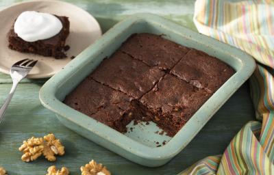 Fotografia em tons de verde em uma bancada de madeira verde, um recipiente verde quadrado com o brownie de chocolate com nozes e um pano colorido ao lado. Ao fundo, um prato redondo bege com uma fatia do brownie.