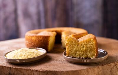 Foto da receita de bolo de milho de liquidificador, redondo, dourado, com uma fatia mais à frente em um prato e ao lado um punhado de fubá em outro prato. Ao fundo é possível ver o bolo inteiro