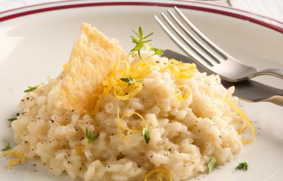 Fotografia de um prato branco com uma listra vermelha na borda, onde está servido o risoto decorando com raspas de limão siciliano e uma crosta crocante de queijo parmesão. Ao fundo uma taça de vidro transparente e um guardanapo de tecido branco.