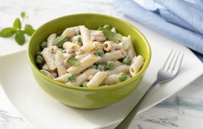 Fotografia em tons de branco, verde e azul de uma bancada branca e um prato retangular branco, sobre ele um recipiente redondo verde com o macarrão e um garfo. Ao fundo um paninho azul e folhas de manjericão.