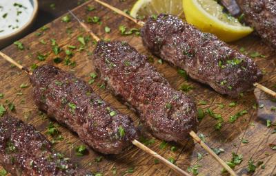 Fotografia de quatro espetinho de carne moída sobre uma tábua de madeira, e por cima, salsinha salpicada. Ao fundo, um molho de pepino dentro de um recipiente de fundo e pequeno, e ao lado, dois pedaços de limão siciliano.