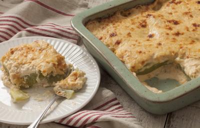 Fotografia em tons de branco e dourado, forma com Chuchu Gratinado sobre bancada de madeira rústica e prato branco com um garfo e uma porção do Chuchu Gratinado sobre guardanapo branco com listras de cor vinho.