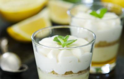 Fotografia de dois copinhos de vidro com farofa de biscoito e creme de limão sobre uma mesa de mármore preta. Ao fundo, duas metades de um limão siciliano, muitas folhas de hortelã e uma colher de sobremesa.