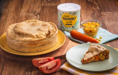 Fotografia em tons de amarelo e verde de uma bancada de madeira, sobre ela um prato amarelo com uma torta, a frente um prato verde com uma fatia da torta, mostrando o recheio de milho, cenoura e tomate. Ao fundo uma lata de Ninho Origem Vegetal.