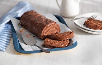 Fotografia em tons de azul, branco e marrom, ao centro da foto o rocambole todo de chocolate com 2 fatias na bandeja azul, ao lado está um prato branco com uma fatia do rocambole e do outro um guardanapo listrado de azul e branco.