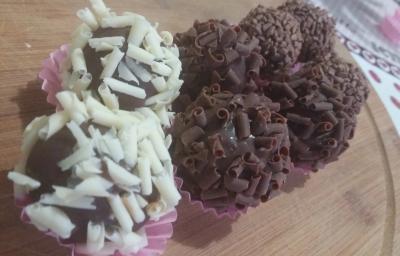 Fotografia de oito brigadeiros dentro de forminhas rosa. Seis brigadeiros foram enrolados em confeitos de chocolate preto, e dois deles foram enrolados em confeitos brancos. As forminhas estão sobre uma forma de madeira.