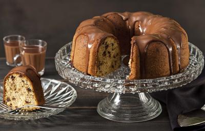 Fotografia em tons de marrom e preto de uma bancada preta vista de frente. Contém um prato redondo transparente com uma fatia de bolo com calda de chocolate por cima, um recipiente transparente para servir o bolo com o bolo sem uma fatia e calda por cima.