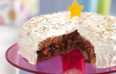 Fotografia em tons de rosa em uma bancada cinza com um suporte para bolo central na cor rosa com o bolo de aniversário confeitado em cima com cobertura branca e confeitos coloridos, cortado ao meio e recheado com chocolate e morango.