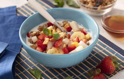 Fotografia em tons de vermelho em uma bancada de madeira de cor branca. Ao centro, um pano azul contendo um bowl azul com as frutas. Ao lado, há um pano azul e ao fundo, um pote com mel e granola.