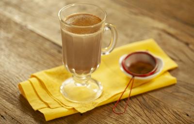 Fotografia em tons de marrom e amarelo de uma bancada de madeira com um paninho amarelo, sobre ele uma xícara transparente com leite, chocolate baton derretido e canela em pó. Ao lado uma peneira vermelha com canela.