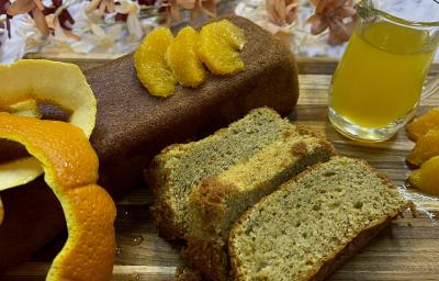 Foto da receita de Bolo de Laranja com Amêndoas. Observa-se um bolo inglês com três pedaços cortados. Como decoração, suco de laranja e a casca cortada.