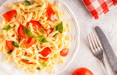 fotografia em tons de branco, amarelo e vermelho de uma bancada branca vista de cima. Contém um prato redondo branco com macarrão tipo talharim com pedaços de tomate e folhas de manjericão. Ao lado garfo e faca para servir.