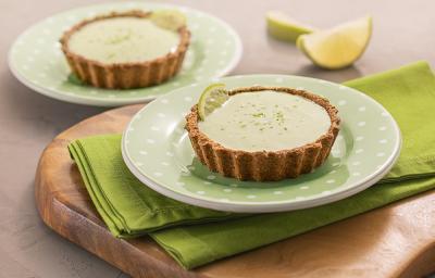 Foto da receita de tortinha de limão. Observa-se dois pratos verdes de sobremesa com as tortinhas sobre um guardanapo verde e uma tábua de madeira. Atrás, duas fatias de limão.