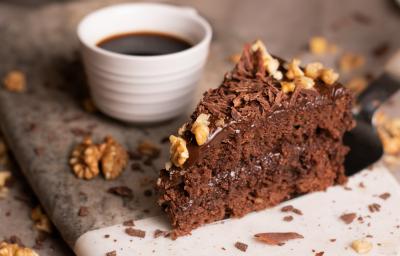 Fotografia de uma fatia de bolo feito com Chocolate NESTLÉ CLASSIC Meio Amargo, nozes e Cobertura de Chocolate ao Leite GAROTO, com raspas de chocolate e castanhas por cima. Ao lado da fatia tem uma xícara pequena branca com café.