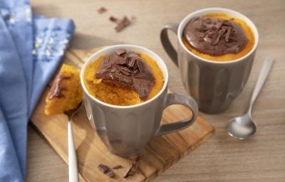 Fotografia em tons de laranja em uma bancada de madeira clara, uma tábua de madeira, um paninho azul, duas canecas marrons com o bolo de cenoura dentro de cada uma e por cima o brigadeiro de Nescau.