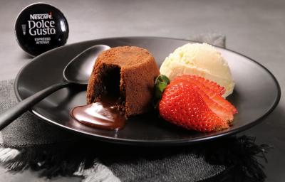 Fotografia em tons de preto em uma bancada de madeira escura, um prato preto redondo com o petit gateau de café em cima, ao lado, uma bola de sorvete de creme e um morango para decorar. Ao fundo, uma cápsula do café intenso de Dolce Gusto.