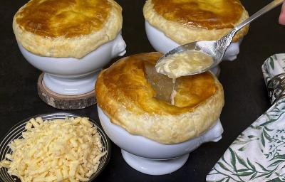 Foto em tons de escuros da receita de sopa de cebola en croute servida em três porções sobre bases de porcelana branca, sendo que há uma colher prateada pegando um pedaço da porção da frente. Ao lado há um prato pequeno com queijo emmenthal ralado